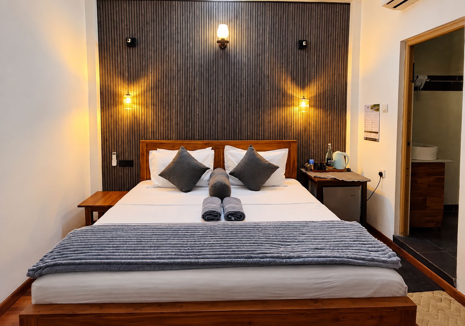A neatly made bed with pillows, a wooden headboard, and a nightstand with a lamp and a bottle.