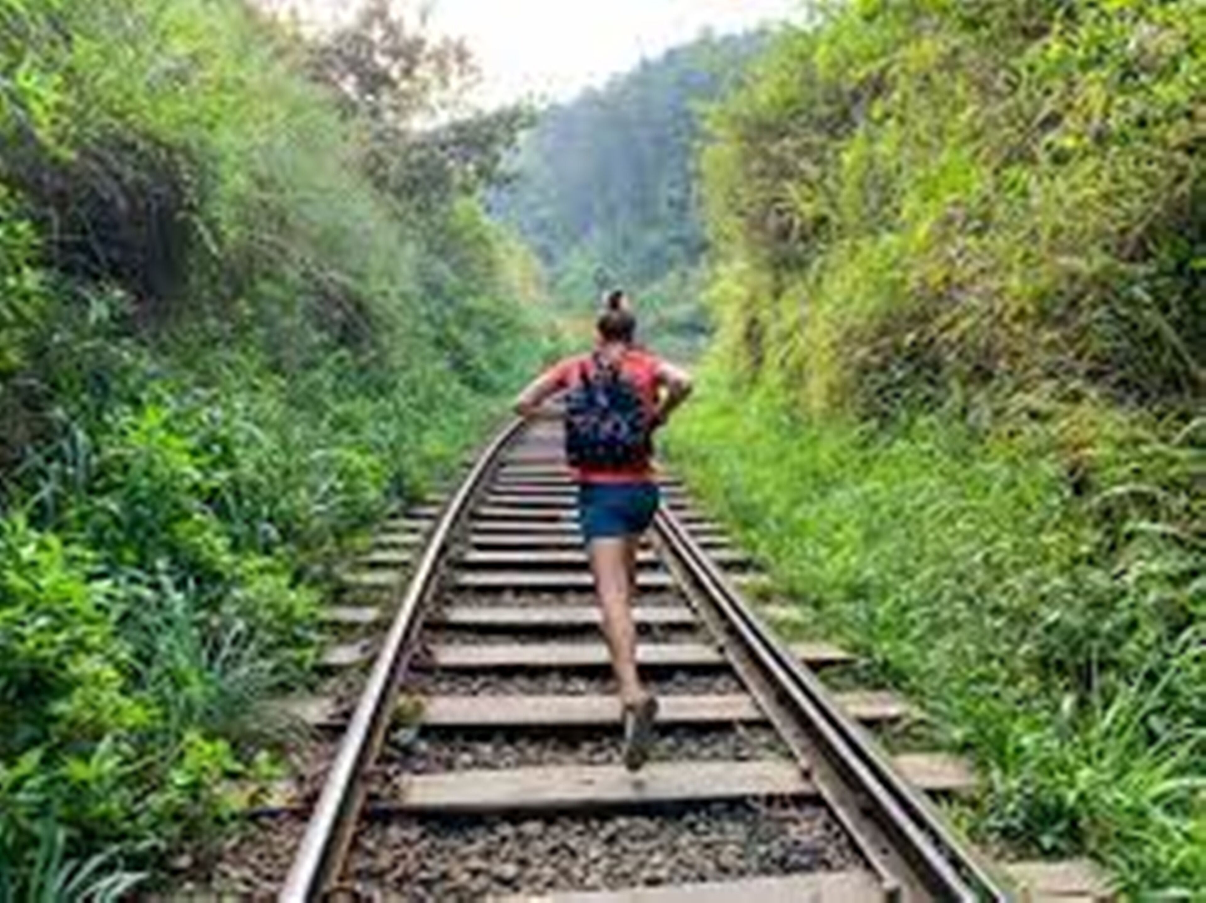 Una persona caminando sobre una vía de tren rodeada de vegetación.