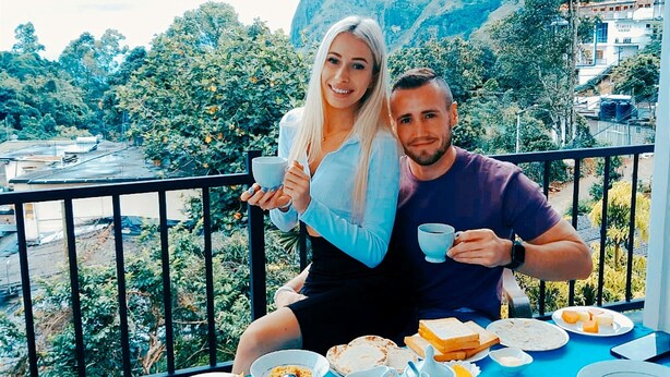 Un couple prend son repas sur un balcon, une table est dressée pour le petit-déjeuner, et la vue donne sur un paysage montagneux.