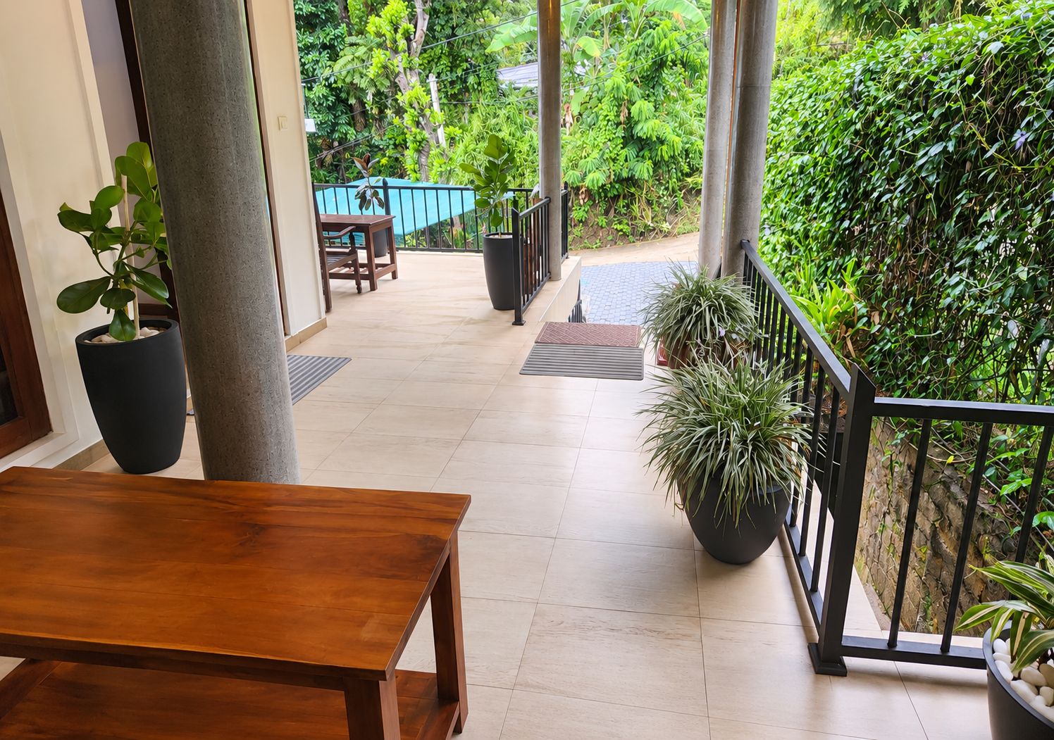 Una mesa y sillas de madera en un balcón con plantas en macetas, barandal azul y vistas de vegetación exuberante.