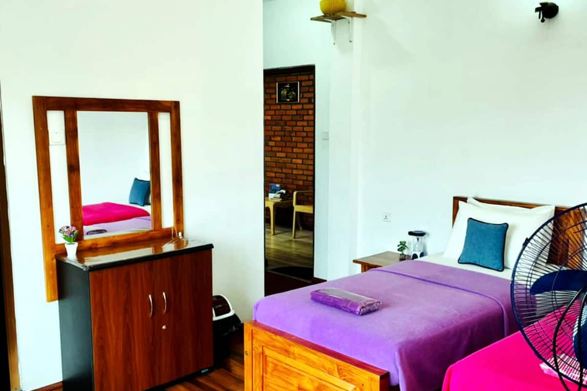 Une chambre avec une armoire en bois, un lit avec une literie violette, une petite table avec un ventilateur et un mur en briques en arrière-plan.