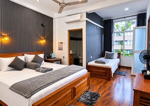 A hotel room with a large bed, a smaller bed, a desk, a chair, a fan, and a window with a view of greenery.