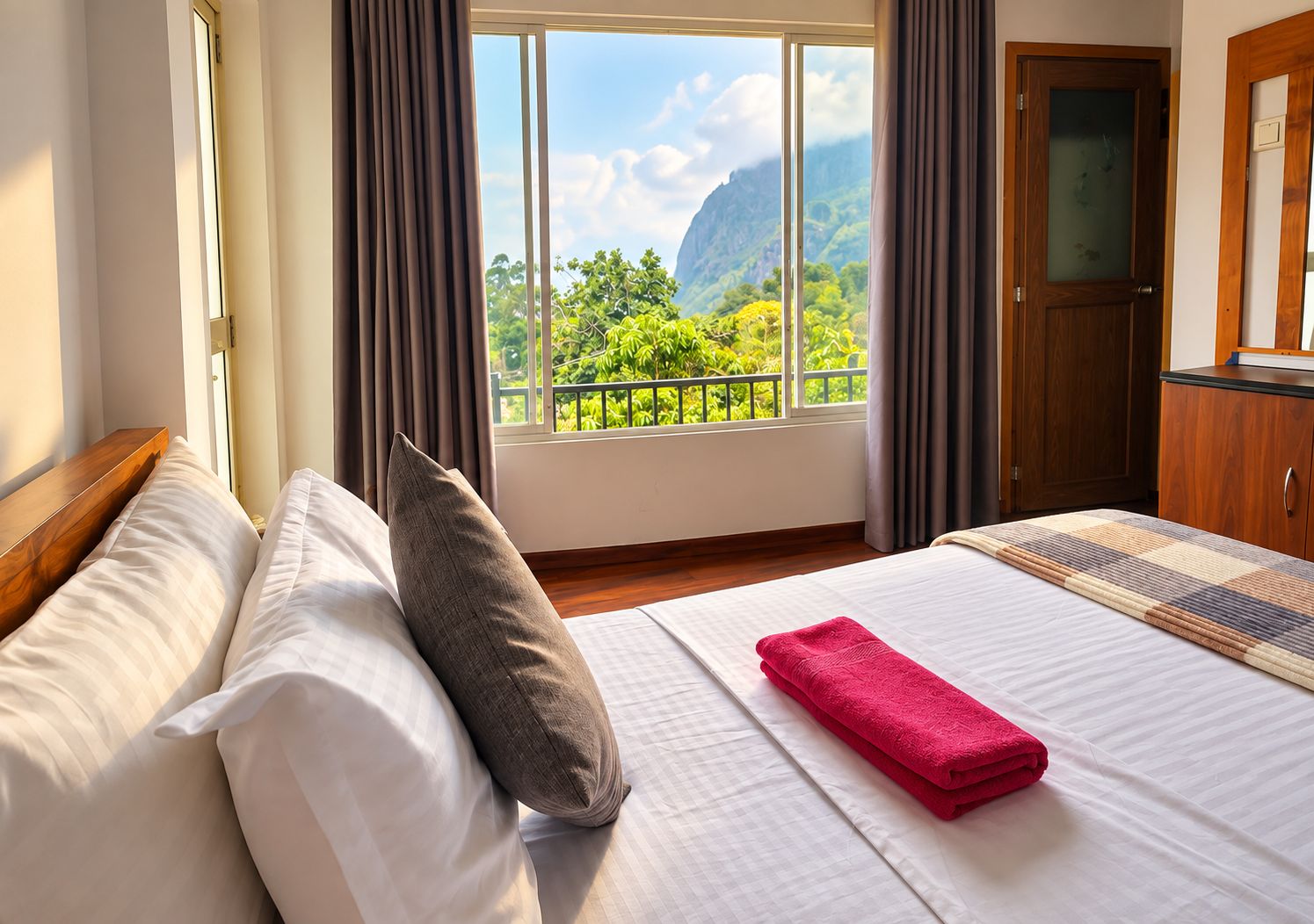 Una habitación de hotel con cama, ventana con cortinas, vistas a zonas verdes y montañas, y una puerta de madera.