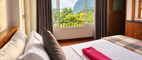 Una habitación de hotel con cama, ventana con cortinas, vistas a zonas verdes y montañas, y una puerta de madera.