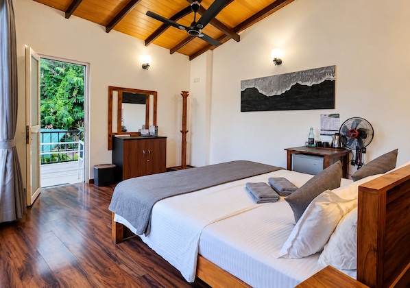 A bedroom with a bed, a dresser, a desk, a fan, and a view of greenery outside.