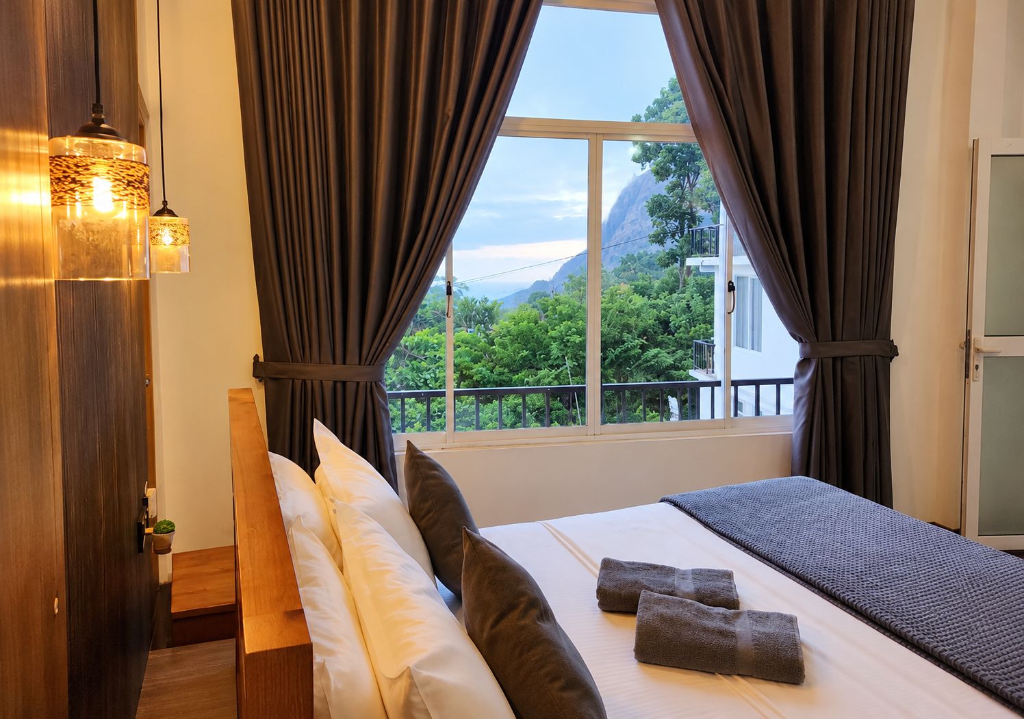 A hotel room with a bed, pillows, a view of greenery and mountains, and a window with curtains.