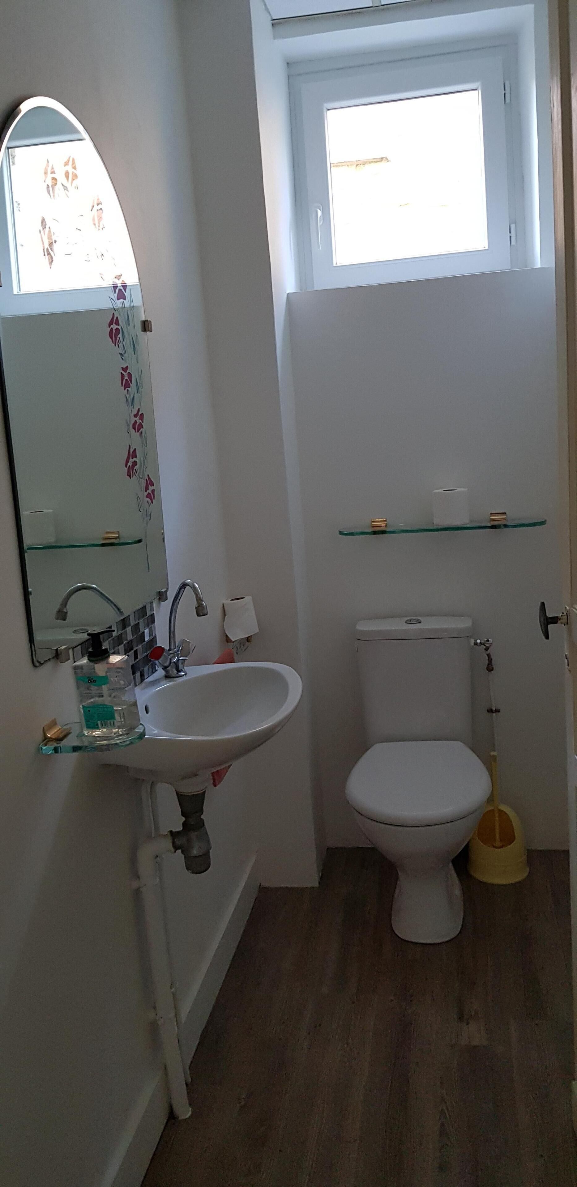 Une petite salle de bain avec un lavabo blanc, des toilettes, un miroir et une fenêtre située au-dessus des toilettes.