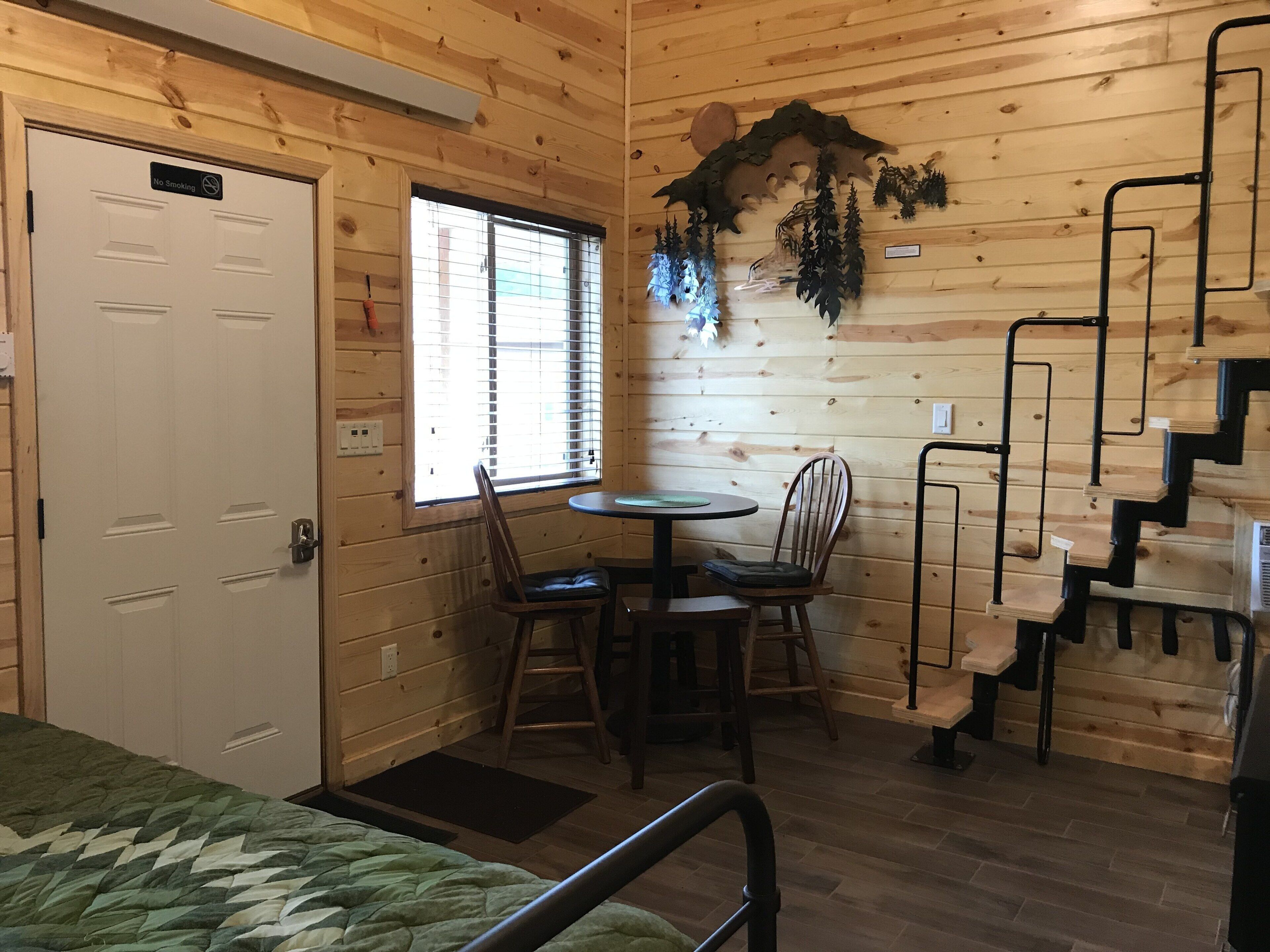 L’intérieur d’un chalet en bois comprend un lit, une table à manger avec des chaises, une fenêtre avec des stores, un escalier et un panneau indiquant qu’il est interdit de fumer.
