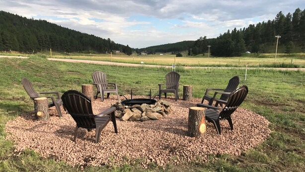 A fire pit with four Adirondack chairs arranged around it, set on a gravel area with a grassy field and forested hills in the background.