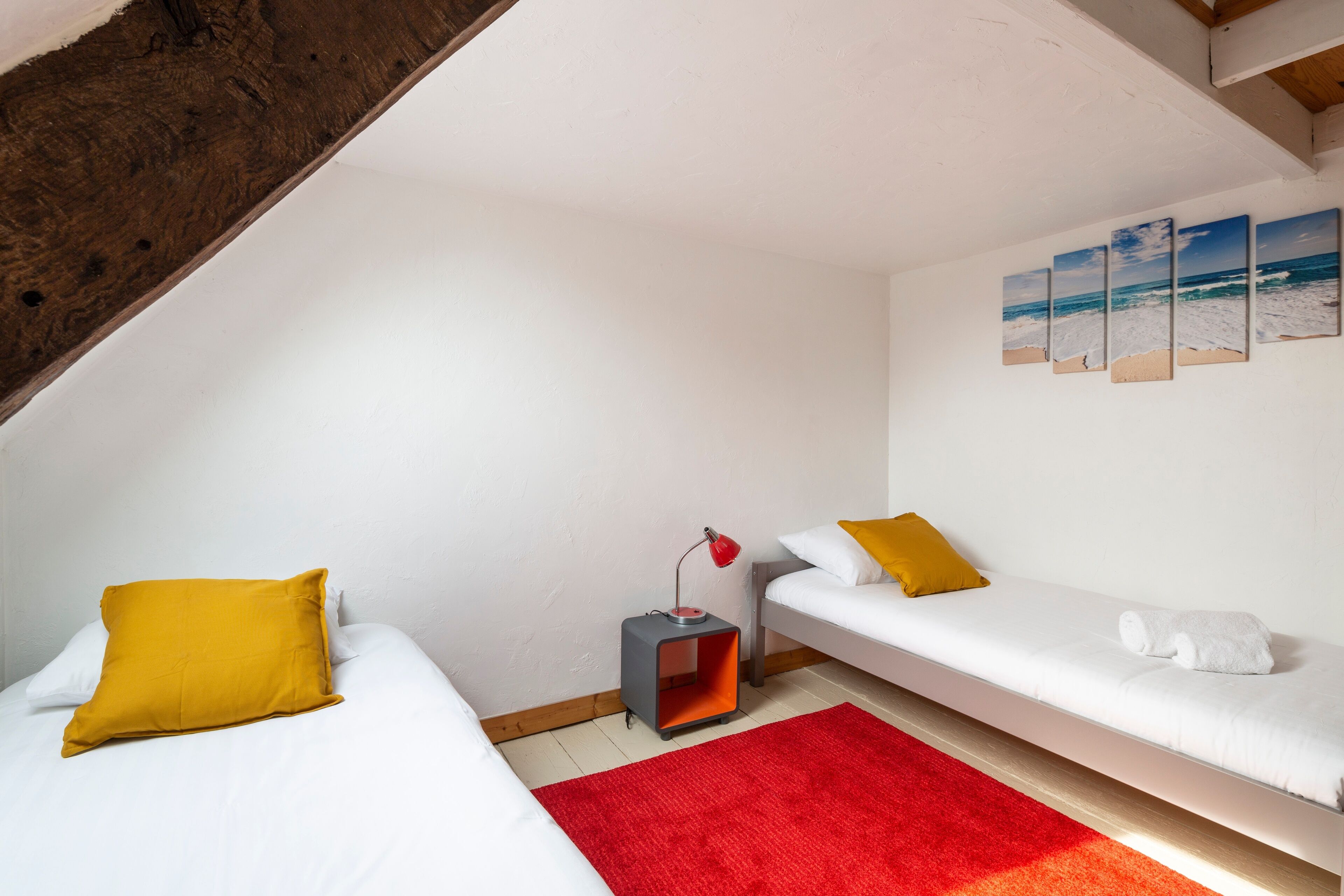 Una habitación con dos camas individuales, un tapete rojo, una mesita con una lámpara y una pared decorada con tres cuadros de temática marina.
