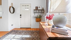 Ein Flur mit einer weißen Tür, Holzboden, einem gemusterten Teppich, einem Holztisch mit Vase und Büchern, einem Wandkorb und einem großen Fenster mit Jalousien.