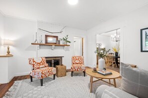 Een woonkamer met een witte bakstenen open haard, twee gestreept fauteuils, een ronde houten salontafel en een grijze bank.