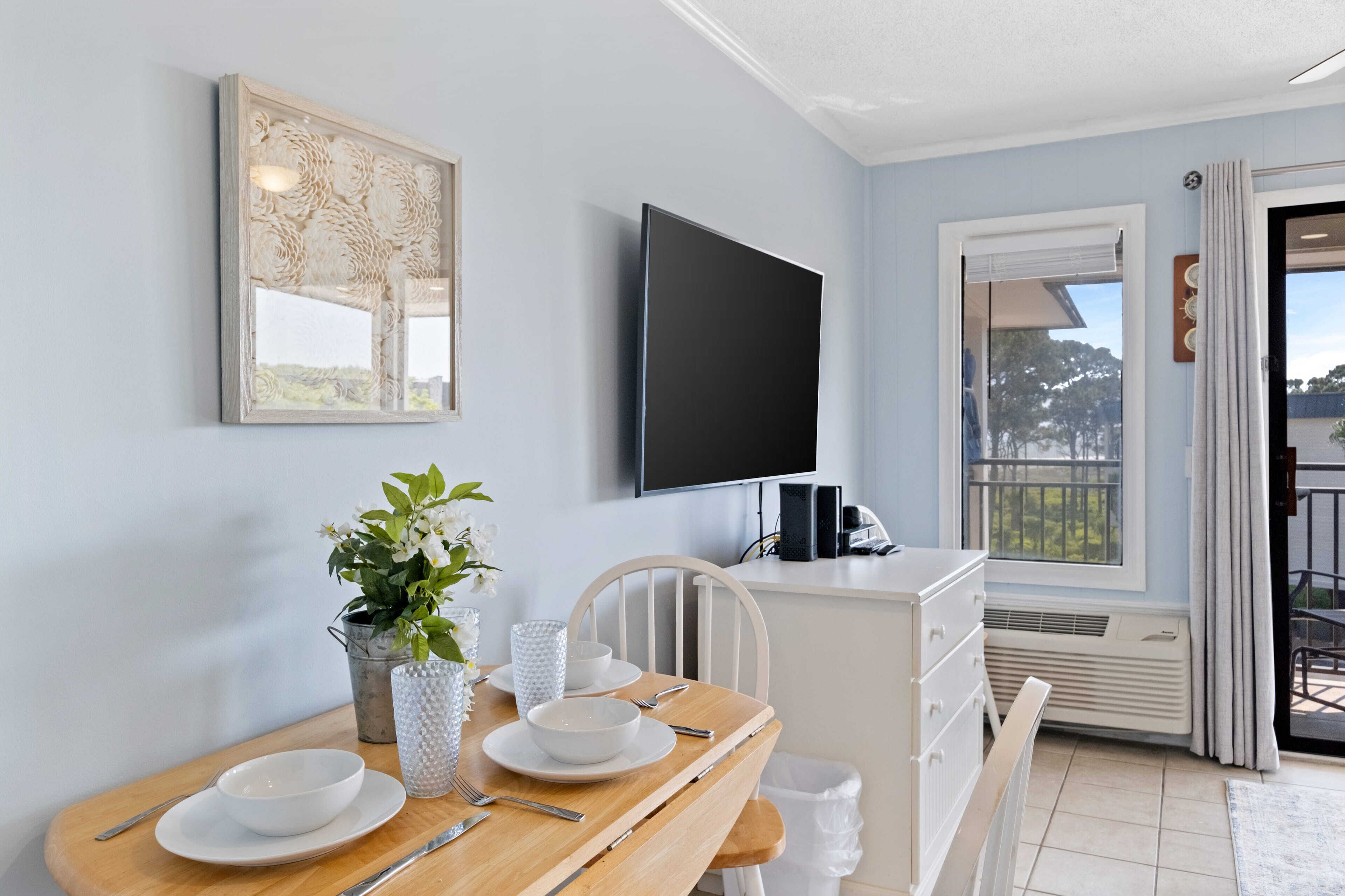 Un comedor con una mesa de madera dispuesta para dos personas, una silla blanca, una planta en maceta y un televisor de pantalla plana montado en la pared.