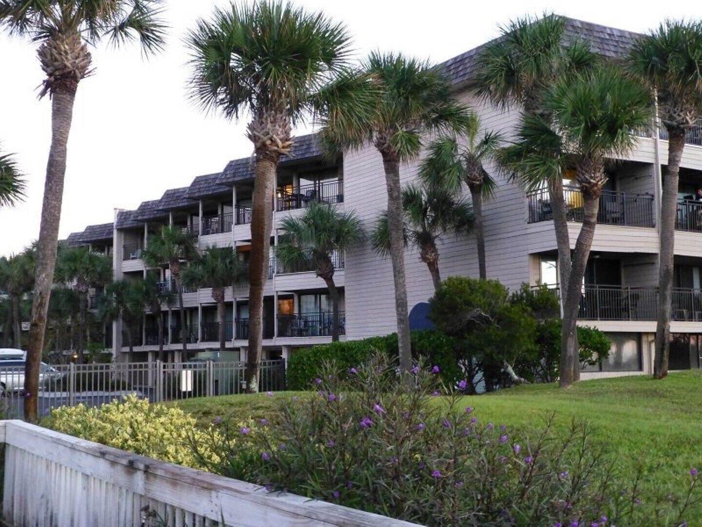 A multi-story hotel with balconies, surrounded by palm trees and a grassy area with flowers.