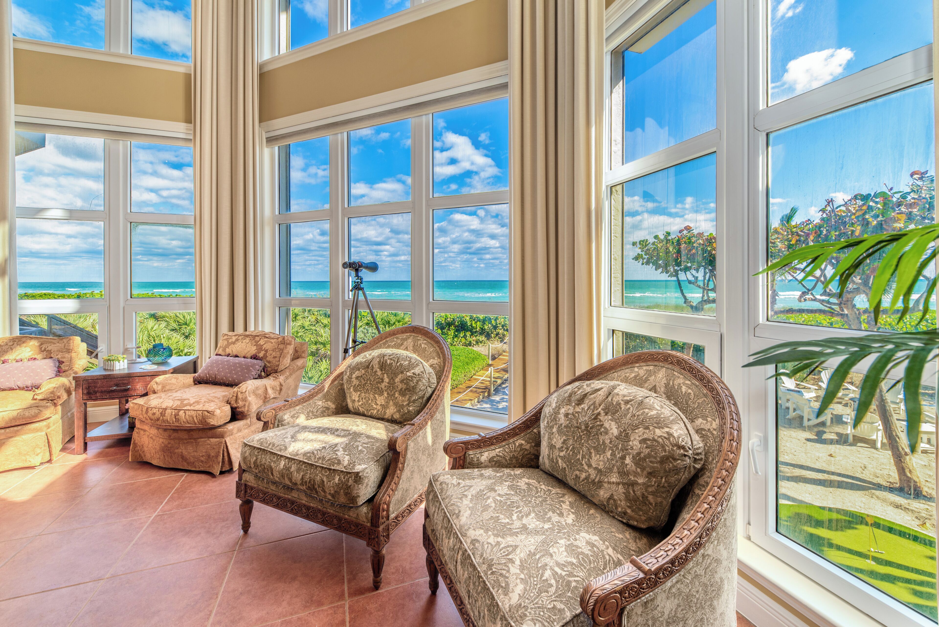 A spacious room with large windows offering a view of the ocean and a golf course. There are several upholstered chairs and a small table.
