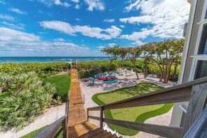 A view from a balcony overlooking a sandy beach area with a golf course, a wooden deck, and a pool with lounge chairs.