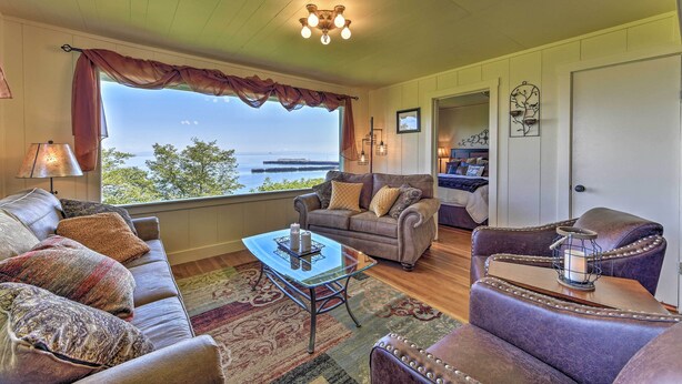 A cozy living room with a large window, a leather sofa, a glass coffee table, and a view of the outdoors. - Waterfront Home ~ 6 Mi to Olympic National Park! (Port Angeles)