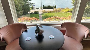 Habitación con vista a un jardín ajardinado y un cuerpo de agua a través de grandes ventanales. Cuenta con una mesa redonda con dos sillas y una pequeña figurita sobre la mesa.