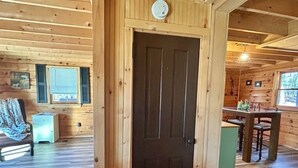 A wooden cabin interior with a dark wooden door, a bed with a blanket, a small table with chairs, and a window with blinds. - Hidden Gem in the woods with hot tub  (Berkeley Springs)