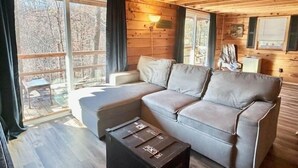 A cozy cabin interior with a sectional sofa, a small table, and a large window offering a view of the outdoors. - Hidden Gem in the woods with hot tub  (Berkeley Springs)
