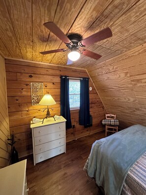 Ein gemütliches Schlafzimmer im Dachgeschoss mit Holzwänden, einem Deckenventilator, einem Fenster mit Vorhängen, einem Nachttisch mit Lampe, einer Kommode und einem Bett.