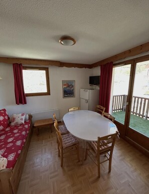 A cozy room with a wooden floor, a round dining table with chairs, a sofa with red cushions, a radiator, and a window with curtains.