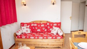 Una habitación acogedora con un sofá cama de madera cubierto con ropa de cama estampada en rojo y blanco, dos lámparas fijadas a la pared, una mesa de madera con un mantel individual y una cortina roja.