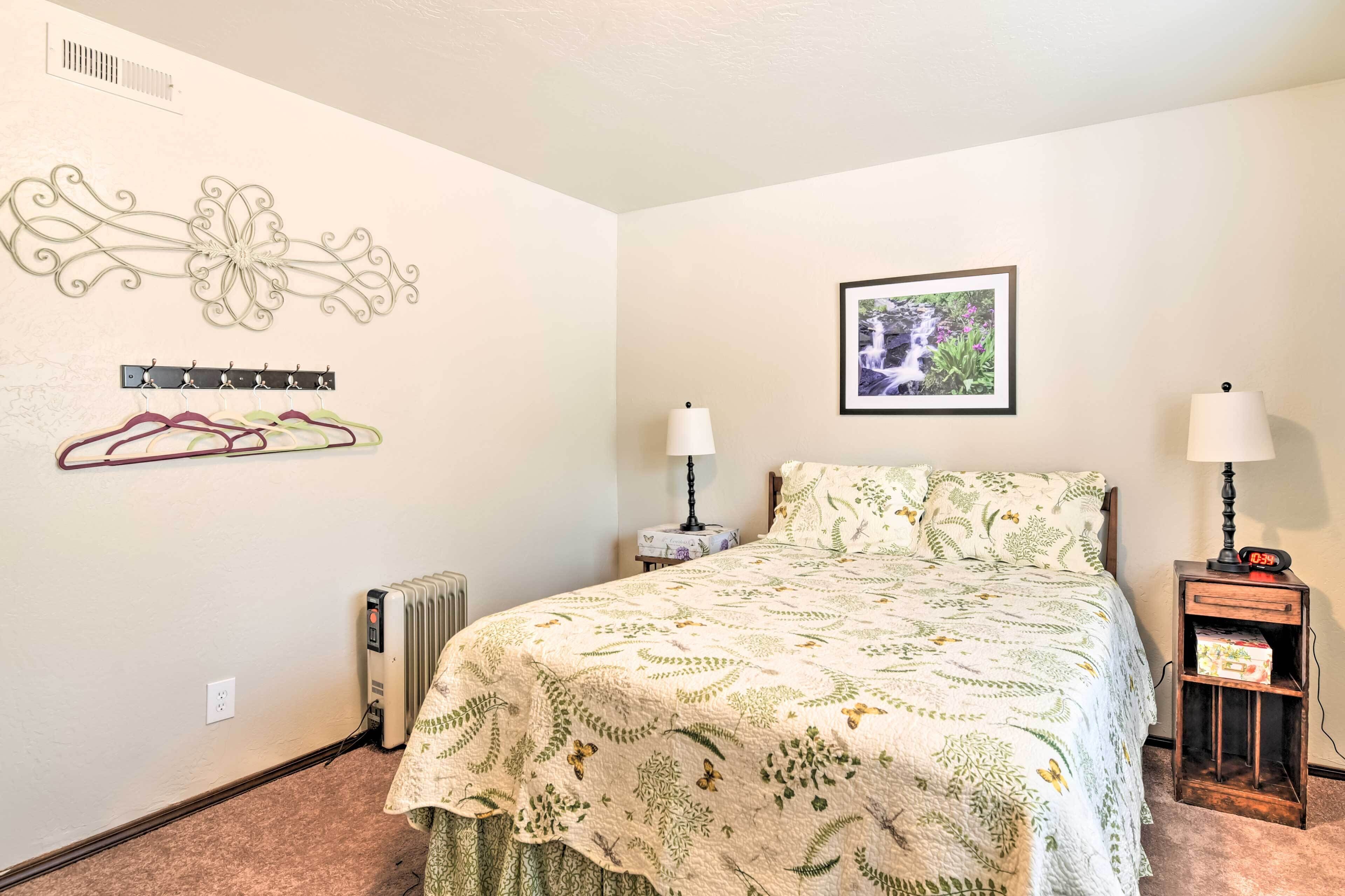 A bedroom with a bed, two bedside tables with lamps, a radiator, a wall-mounted coat rack, a framed picture, and a decorative wall piece.