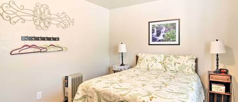 A bedroom with a bed, two bedside tables with lamps, a radiator, a wall-mounted coat rack, a framed picture, and a decorative wall piece.