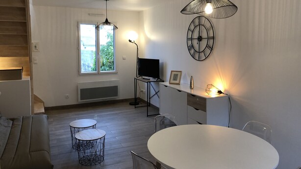 Ein modernes Wohnzimmer mit einem weißen Sofa, einem runden Esstisch und zwei kleinen Beistelltischen. Ein Fernseher steht auf einem Schrank, eine Uhr hängt an der Wand und ein Fenster bietet Blick ins Grüne.