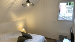 Ein Schlafzimmer mit einem Bett, einer Nachttischlampe, einem kleinen Tisch mit Fernbedienung und einem Fenster mit Blick auf Gebäude.