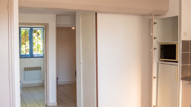 Une porte ouverte donne sur une pièce dont le sol est recouvert de carreaux, une fenêtre ornée d’un cadre bleu, et un four à micro-ondes posé sur un meuble.