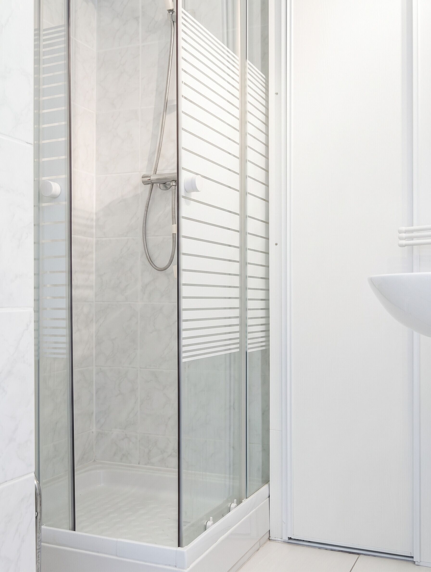 Eine gläserne Dusche mit Duschbrause, eine weiß geflieste Wand und ein Waschbecken mit gebogener Spüle.