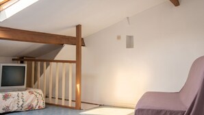 Une pièce avec un plafond en pente, des poutres en bois, un téléviseur, un fauteuil à motifs floraux et une petite fenêtre.