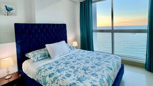 Un dormitorio con una cama grande, una mesita de noche, una lámpara y vistas al mar a través de un ventanal amplio.