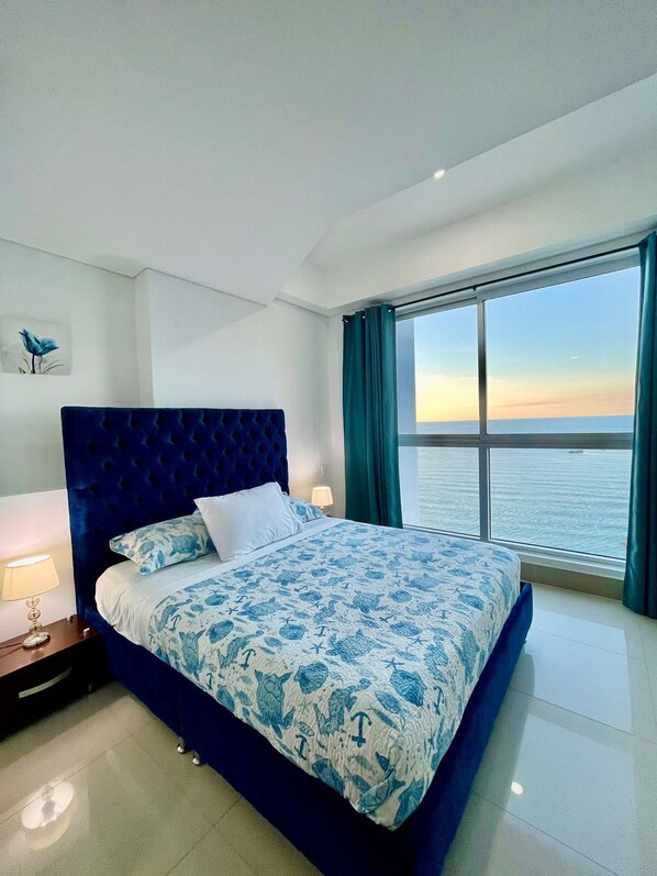A bedroom with a large bed, a nightstand, a lamp, and a view of the sea through a large window.