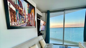 Una sala moderna con un ventanal que ofrece vista al mar, un sofá color beige, un televisor de pantalla plana y un cuadro de una escena costera.