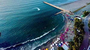 Vista aérea de una zona costera con una playa, un muelle que se adentra en el mar y un paisaje urbano distante.