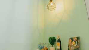 Um quarto com uma luminária pendente geométrica, um pequeno vaso com planta, uma garrafa de vinho, um quadro emoldurado e um recipiente de vidro decorativo sobre uma mesa de madeira.