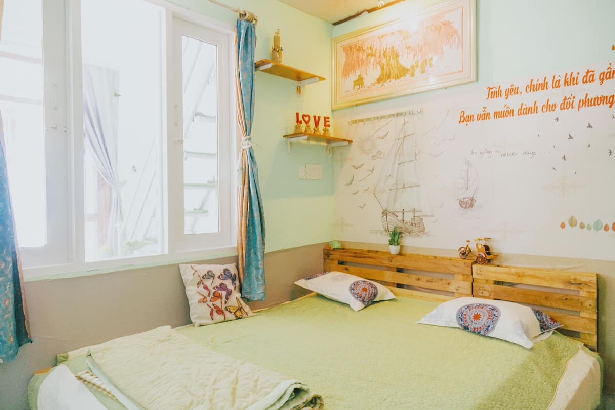 A bedroom with a bed, pillows, a window with curtains, a wall with a map and a 'LOVE' sign, and a small shelf with decorative items.