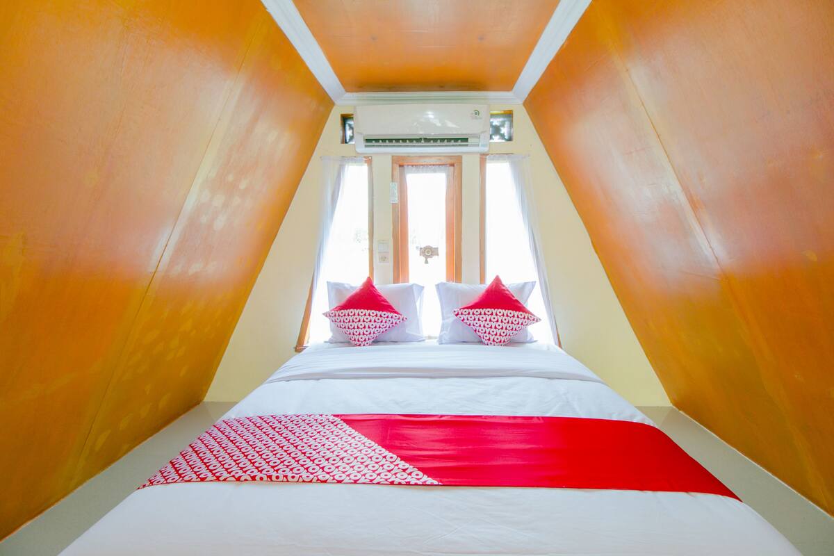 A room with a sloped ceiling, a bed with white bedding and red accents, and a window with curtains.