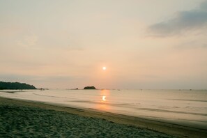 Une plage avec une mer calme, un soleil bien visible et une terre ferme qui se dessine à l’horizon.