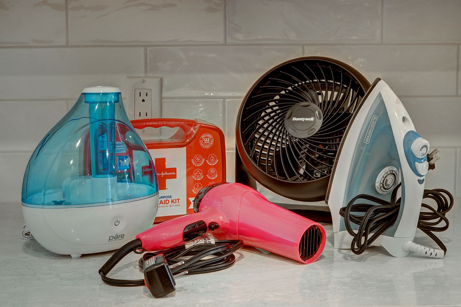 Une cuisine avec un mur recouvert de carreaux blancs, un distributeur d’eau bleu, une trousse de premiers secours rouge, un ventilateur noir de la marque Honeywell, un sèche-cheveux rose et un fer à repasser électrique.