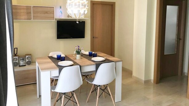 A dining area with a wooden table, white chairs, a flat-screen TV mounted on the wall, and a decorative globe light.