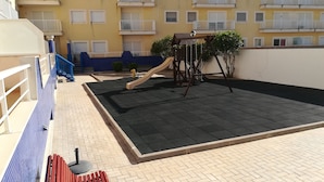 A rooftop terrace with a playground, including a slide and swings, surrounded by a low wall and a blue railing.