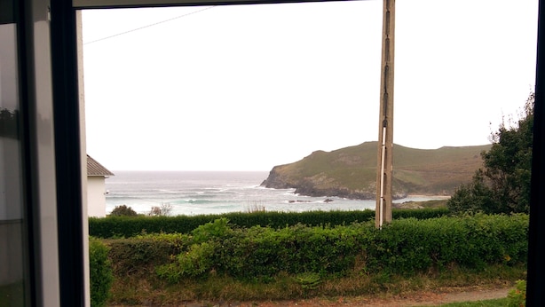 Vista a través de una ventana que muestra un área verde con un sendero, un seto y un paisaje costero que incluye el océano y colinas.