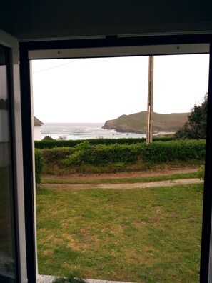 Vista a través de una ventana que muestra un área verde con un sendero, un seto y un paisaje costero que incluye el océano y colinas.