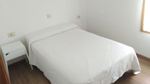 Una cama pequeña y ordenada con una colcha blanca en una habitación minimalista con paredes blancas y suelo de madera.