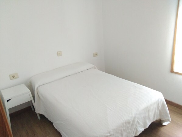 Una cama pequeña y ordenada con una colcha blanca en una habitación minimalista con paredes blancas y suelo de madera.