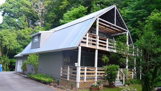 Ein zweigeschossiges Haus mit einem geneigten Metalldach, einer Veranda mit Holzgeländer und einem kleinen Gartenbereich mit Blumentöpfen.